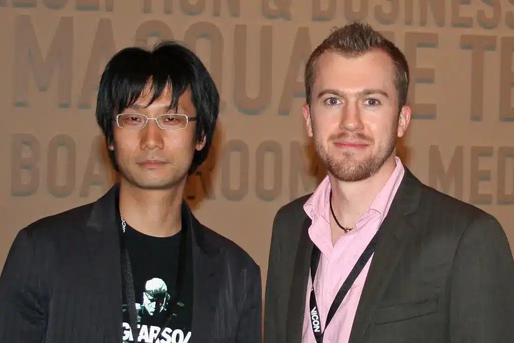 眼鏡をかけ黒のブレザーを着たHideo Kojimaが、別の男性の隣に立っている姿。